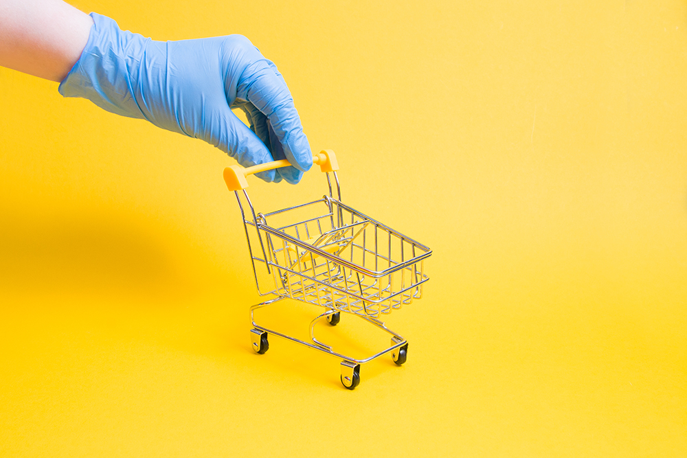 An hand wearing a disposable glove pushes a tiny shopping cart.